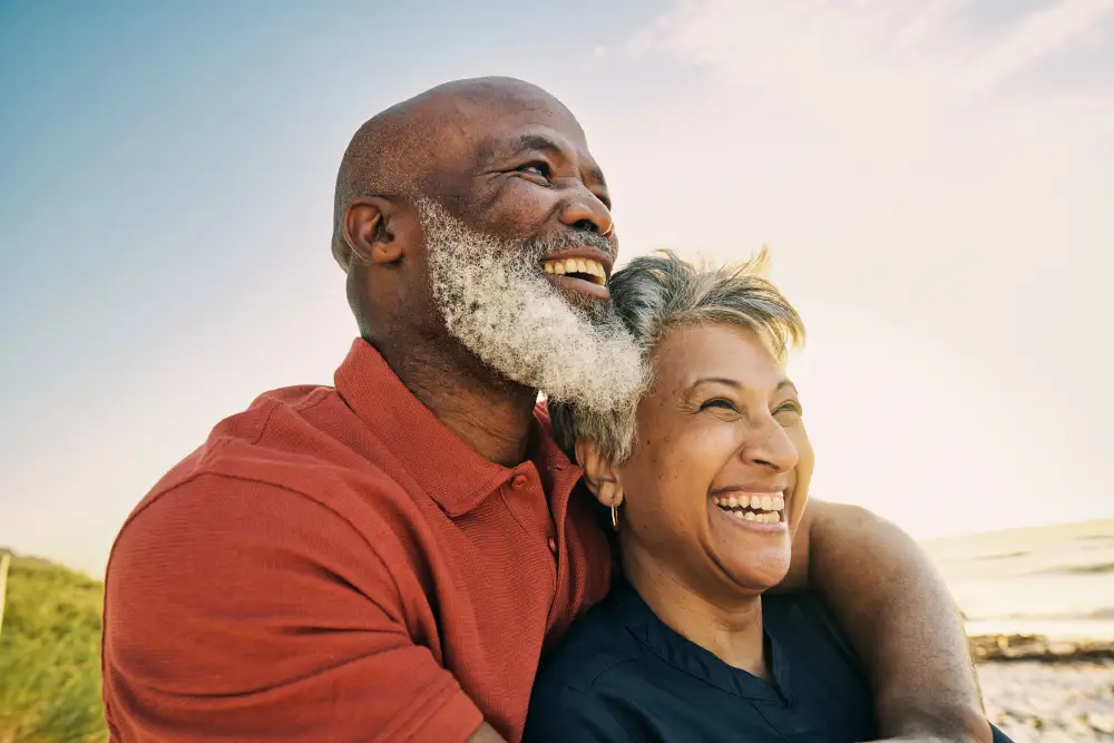 a senior couple smiling while standing on the beach and watching the sun set